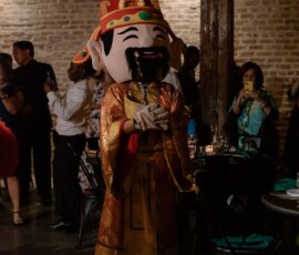 Person in Chinese deity costume at a festive, dimly lit brick-walled event, possibly a lively Gulf South wedding.