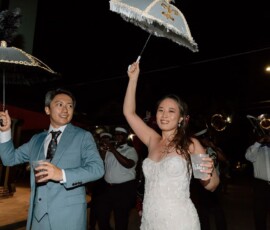 Bride and groom smile under decorated umbrellas at night, with a brass band playing behind at a joyful New Orleans wedding.