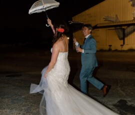 Bride in white gown leads groom in blue suit under a decorative umbrella at night, with a yellow building behind them.
