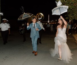 Bride in white dress and groom in blue suit lead lively New Orleans wedding parade at night with musicians and umbrellas.