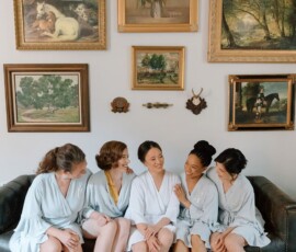Five women in matching robes relax on a couch, smiling and chatting in front of animal and floral art—New Orleans wedding vibes.