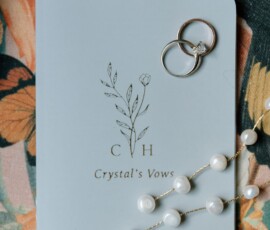 Pale blue “Crystals Vows” booklet with rings and pearl necklace on floral fabric—ideal for a New Orleans wedding.