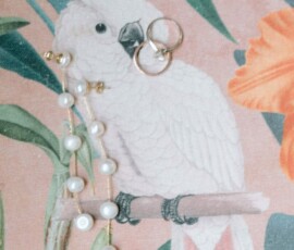Gold rings and a pearl necklace rest on fabric featuring a white parrot among tropical flowers and green leaves.