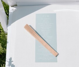 Light blue wedding program and wooden stick on a white outdoor chair, with grass and ferns nearby at a Louisiana wedding.