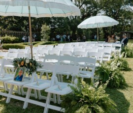 Outdoor New Orleans wedding with rows of white chairs, two large umbrellas, greenery, and guests standing under trees.