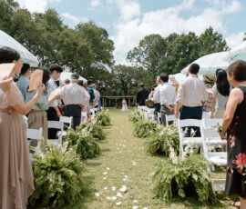 Guests watch a bride in white walk down a flower-petal aisle toward the altar, surrounded by trees and ferns under sunny skies.