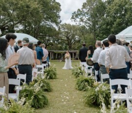 Bride walks down an outdoor aisle with white chairs and greenery at a New Orleans wedding as guests watch under a cloudy sky.