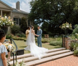 Bride and groom hold hands outdoors on steps, bridesmaid with flowers nearby, large building and greenery in background.