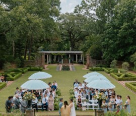 Outdoor garden wedding with guests under blue umbrellas, a couple at the altar, lush greenery, and sunny Gulf South vibes.