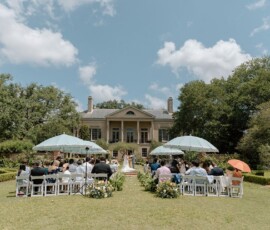 Sunny outdoor Louisiana wedding with guests under umbrellas, couple at altar, elegant house, and lush greenery.