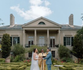 Bride and groom hold hands at an outdoor Louisiana wedding before a grand mansion, with a gold-clad officiant between them.