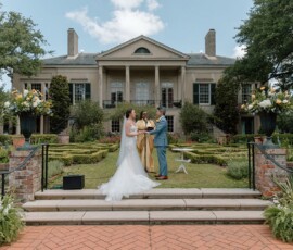 Bride and groom with officiant in front of elegant mansion, lush gardens at sunny Louisiana outdoor wedding.