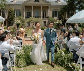 Bride and groom smile walking down an outdoor aisle, guests seated near a grand mansion and lush gardens on a sunny day.