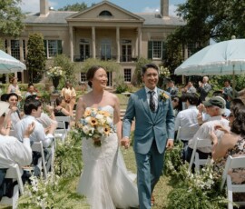 Smiling bride and groom walk hand in hand down the aisle at an outdoor Gulf South wedding by a large house.