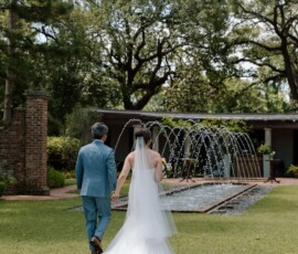 Bride in white gown and groom in blue suit walk toward a fountain on a Louisiana lawn, framed by trees and a garden building.