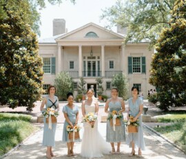 Five women in pastel blue dresses hold flowers outside a columned mansion, classic Louisiana wedding setting, sunny day.