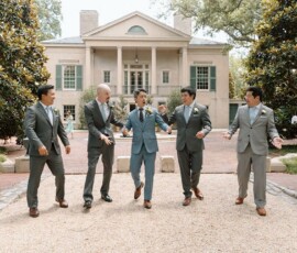 Five men in suits, one in light blue, laugh as they walk a gravel path by a grand house with columns and greenery.