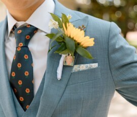 Person in a light blue suit with sunflower boutonniere and tie, plus photo pin. Sunlit, blurred wedding-inspired background.
