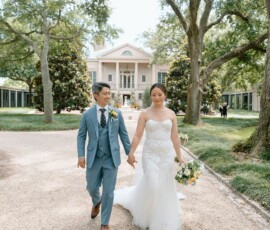 Bride in white dress and groom in blue suit walk hand in hand on a path, smiling by trees and a mansion at a Louisiana wedding.