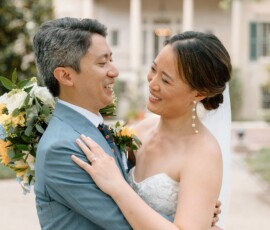 Bride in white dress and groom in blue suit embrace outside a historic Louisiana building, smiling with bouquet.