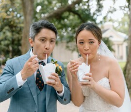 Bride and groom in wedding attire sip from white cups with straws, smiling outdoors among Louisiana greenery.