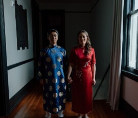 Couple stands together in a dim hallway before their New Orleans wedding, blue robe and red dress catching soft window light.