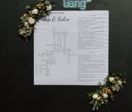 Sip & Solve crossword on a black wall with flowers, neon #ZhengGang sign, and markers—stylish Gulf South wedding inspo.