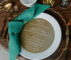 Elegant table setting with gold cutlery, green napkin, and Crystal & Hieu’s May 13, 2023 Gulf South wedding menu.