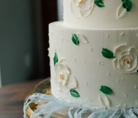 White tiered cake with white flowers and green leaves on a glass stand, yellow flowers and blue ribbon at the base.