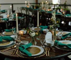Elegant wedding table with gold plates, green napkins, white flowers, candles, and matching decor in the background.