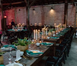 Elegant wedding tables with gold cutlery, green napkins, candles, and plants in a rustic Louisiana venue with brick walls.