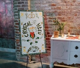 Wedding sign on easel reads “Crystal & Hieu, May 13, 2023” with florals; set by a brick wall near flowers and candle.