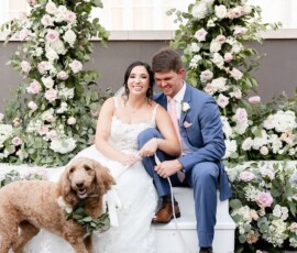 Bride and groom smile by floral arches at their Louisiana wedding, with a golden doodle in a wreath and pink-white blooms.