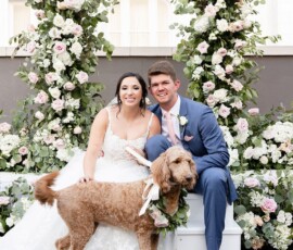Bride and groom smile by a floral arch at their Louisiana wedding, with their brown dog in a flower collar nearby.
