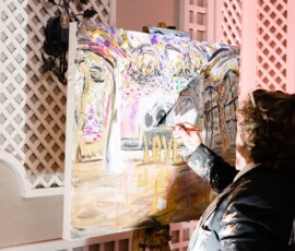 An artist with shoulder-length hair paints a vibrant abstract scene on canvas in a softly lit room with white lattice walls.
