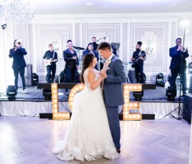 Bride and groom share their first dance in a bright room with a live band and illuminated LOVE letters behind them.