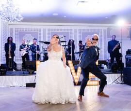 Bride and groom dance playfully before lit LOVE letters and a live band at a bright indoor Gulf South wedding reception.