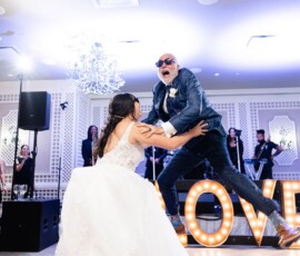 Bride playfully lifts man in sunglasses mid-jump at a New Orleans wedding, with glowing LOVE sign in the background.