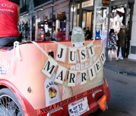 A red pedicab with a Just Married sign drives down a city street at night, surrounded by lit shops after a Louisiana wedding.