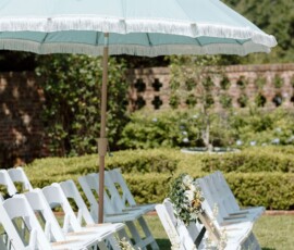 White folding chairs with flowers are set in rows beneath a blue umbrella in a garden with greenery and brick wall backdrop.