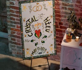 Colorful wedding welcome sign for Crystal ♥ Hieu, May 13, 2023, with flowers and rings beside a white table on a brick wall.