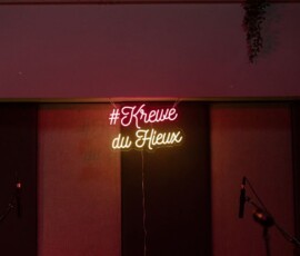 Neon #Krewe du Vieux sign in pink and yellow glows on a dim wall, with two microphones at the bottom.