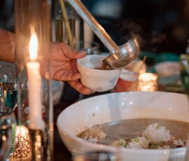 Ladling hot soup into a white bowl at a candle-lit table with glassware, greenery, and a large serving dish.