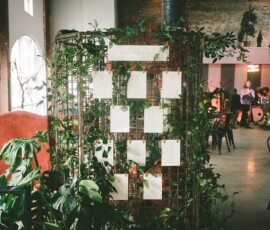Metal grid with greenery displays wedding seating cards in a rustic indoor venue filled with natural light.