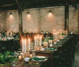 Rustic table with green napkins, gold plates, candles, and plants at a Gulf South wedding in a brick-walled, warmly lit venue.