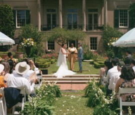 Bride and groom with officiant at outdoor wedding by a mansion, surrounded by guests and greenery in New Orleans.