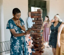 Woman in floral dress removes Jenga block at lively Louisiana wedding as guests watch, some standing and one drinking.