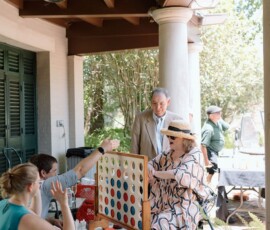 Adults play giant Connect Four at a joyful Louisiana wedding, with guests smiling on a shaded patio amid greenery and sunlight.