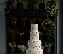 Five-tier white wedding cake with white flowers and greenery on a wooden table, sunflower glasses, and potted plants behind.