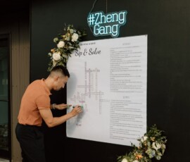 Guest in orange shirt solves large crossword on black wall with flowers and neon #ZhengGang sign at Gulf South wedding.